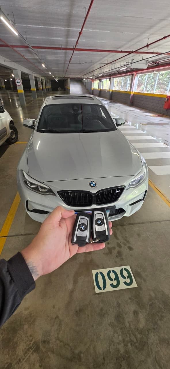 Duas chaves presenciais BMW seguradas em frente a um BMW branco na vaga 099